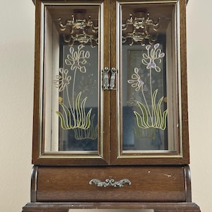 May include: A wooden jewelry armoire featuring two glass doors with painted floral designs in green and purple. The interior contains gold-toned necklace hooks and a mirrored back. A single storage drawer with an ornate metal handle sits below.