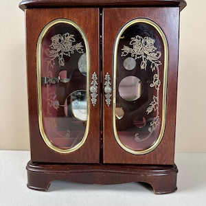 May include: A dark wood jewelry armoire with two doors featuring oval glass panels. Each glass panel is etched with a floral rose design and framed in gold-tone metal. The doors have ornate silver-tone metal handles and a pink interior lining.