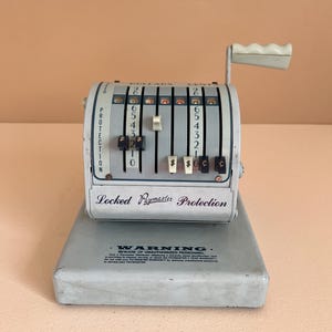 May include: A vintage white cash register with a black handle and a warning label that reads "WARNING. BEWARE OF UNAUTHORIZED PERSONNEL". The register has a black and white dial with numbers 0-9 and a dollar sign.