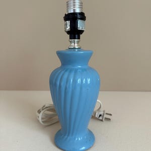 Peut inclure: Petite lampe de table en céramique bleu clair avec un motif en spirale. La lampe est dotée d'une douille noire et argentée et d'un cordon électrique blanc avec une prise à deux broches. La lampe est conçue pour accueillir une ampoule.