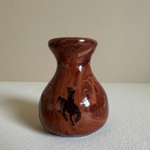 May include: A small, bulbous ceramic vase with a glossy, marbled finish in shades of reddish-brown and tan. The front of the vase features a solid black silhouette graphic of a person riding a bucking horse, commonly known as a bucking bronco.