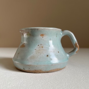 Puede incluir: Una pequeña jarra de cerámica azul claro con un asa marrón. La jarra tiene un esmalte moteado y una forma ligeramente redondeada.