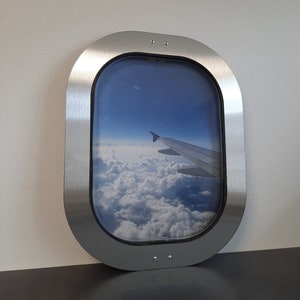May include: A silver airplane window frame with a view of clouds and a wing of an airplane in the distance.