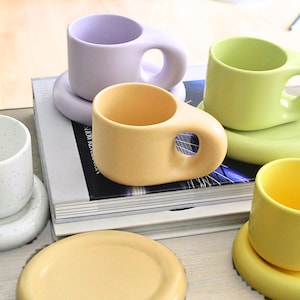 Op de afbeelding: Een verzameling kleurrijke keramische mokken en schotels in verschillende pastelkleuren, waaronder lavendel, geel en groen. De mokken hebben een uniek handvatontwerp met een grote opening. De schotels zijn rond en passen bij de kleur van de mokken.