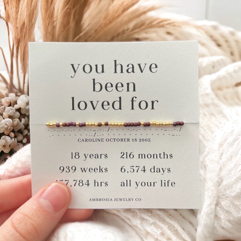 K&ouml;nnte beinhalten: Eine wei&szlig;e Karte mit dem Text "you have been loved for" und einem Armband mit goldenen und burgunderfarbenen Perlen. Die Karte enth&auml;lt auch das Datum "CAROLINE OCTOBER 18 2005" und den Text "18 years, 216 months, 939 weeks, 6,574 days, 7,784 hrs all your life". Die Karte ist signiert mit "AMBROSIA JEWELRY CO".