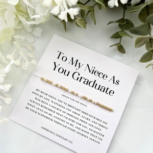 May include: A white card with gold lettering that reads "To My Niece As You Graduate" and a gold and white beaded bracelet. The card has a message for a niece, wishing her well in her future endeavors.  The card is from Ambrosia Jewelry Co.