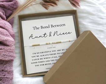 May include: A delicate bracelet featuring white and gold beads arranged in Morse code, presented on a white card inside a brown cardboard box. The card displays the text The Bond Between Aunt & Niece and I LOVE YOU, along with a printed message.