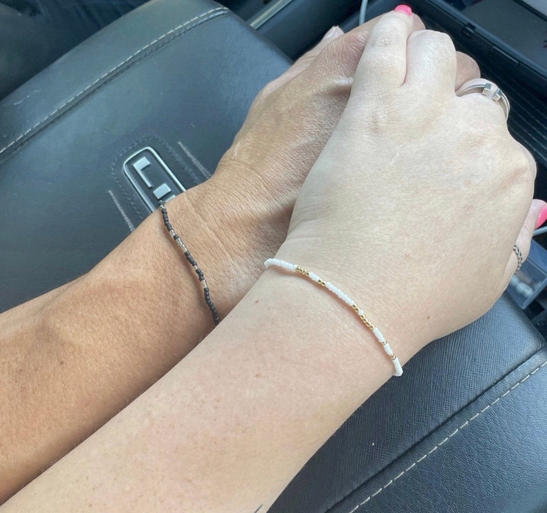 May include: Two people holding hands, one wearing a white beaded bracelet with gold accents and the other wearing a black and white beaded bracelet.