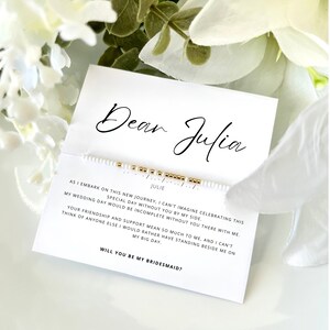 May include: A white card with black text that reads "Dear Julia" and a white beaded bracelet with gold beads. The text on the card reads "As I embark on this new journey, I can't imagine celebrating this special day without you by my side. My wedding day would be incomplete without you there with me. Your friendship and support mean so much to me, and I can't think of anyone else I would rather have standing beside me on my big day. Will you be my bridesmaid?"