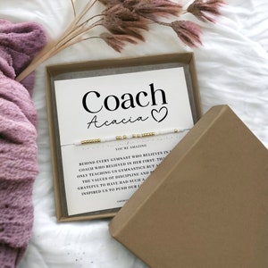May include: A Morse code bracelet with white and gold beads presented on a white card inside a brown cardboard box. The card features the text Coach Acacia with a heart symbol and a message about gymnastics coaching. The bracelet is thin and delicate.