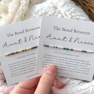 May include: Two cards with the text "The Bond Between Aunt & Niece" and a beaded bracelet on each card. The card on the left has a maroon, gold, and white bracelet and the text "CAROLINE OCTOBER 18". The card on the right has a green, gold, and white bracelet and the text "NATALIE OCTOBER 18 2005".