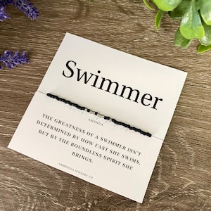 May include: A black and silver beaded bracelet with the text "Swimmer" and a quote about the greatness of a swimmer. The quote reads: "The greatness of a swimmer isn't determined by how fast she swims, but by the boundless spirit she brings." The bracelet also has the word "Amanda" on it and the text "Ambrosia Jewelry Co."