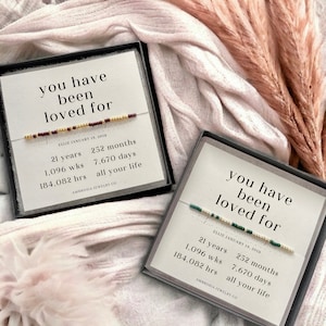 K&ouml;nnte beinhalten: Zwei kleine Schachteln mit schwarzen Deckeln und wei&szlig;en Karten im Inneren. Die Karten tragen den Text "you have been loved for" und ein Datum. Die Karten enthalten auch eine Liste von Zeiteinheiten, darunter Jahre, Monate, Wochen, Tage und Stunden. Die Schachteln befinden sich auf einer rosa-wei&szlig;en, flauschigen Oberfl&auml;che.