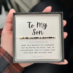 Peut inclure: Un bracelet en perles noires et dorées dans une boîte blanche avec le texte "To my Son" et "I love you unconditionally Alec" sur le devant. La boîte comprend également le texte "Wear this bracelet as a reminder that no matter the place, time, or circumstance, that I'll always have your back and you have mine!" et "Ambrosia Jewelry Co."