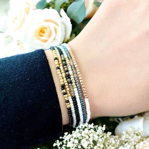 K&ouml;nnte beinhalten: Ein Stapel bunter Perlenarmb&auml;nder am Handgelenk einer Person. Die Armb&auml;nder bestehen aus kleinen, runden Perlen in den Farben Wei&szlig;, Schwarz, Gold und Grau.