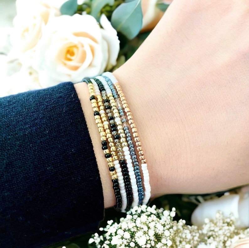 K&ouml;nnte beinhalten: Ein Stapel bunter Perlenarmb&auml;nder am Handgelenk einer Person. Die Armb&auml;nder bestehen aus kleinen, runden Perlen in den Farben Wei&szlig;, Schwarz, Gold und Grau.