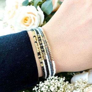 May include: A stack of colorful beaded bracelets in black, white, gold, and silver. The bracelets are layered on a person's wrist.
