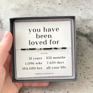 以下が含まれることがあります： 黒い箱の中に白いカードが入っており、黒い文字で「you have been loved for 21 years, 252 months, 1,096 wks, 7,670 days, 184,082 hrs, all your life」と日付「11/11/23」が書かれています。カードは黒い箱に入っていて、下部に「AMBROSIA JEWELRY CO」という文字があります。
