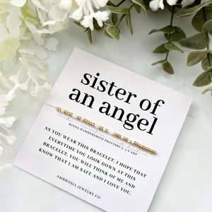 May include: A white card with the text "sister of an angel" and a beaded bracelet. The bracelet has gold and white beads. The card also includes a message and the brand name "Ambrosia Jewelry Co."