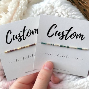 May include: Two cards with the word "Custom" written on them. Each card has a beaded bracelet on it. The bracelet on the left card is made of purple, gold, and silver beads. The bracelet on the right card is made of green, gold, and silver beads.