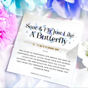 Peut inclure: Bracelet en perles blanches avec des perles dorées et une carte qui dit "Soar & Fly Just Like A Butterfly" avec des illustrations de papillons bleus. La carte comprend également une citation inspirante sur le dépassement des obstacles et un nom d'entreprise, "Ambrosia Jewelry Co."