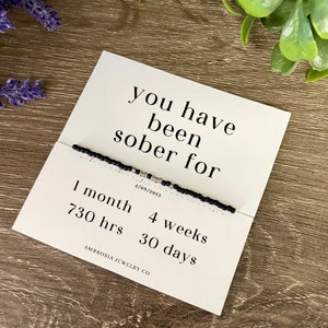 May include: A white card with the text "you have been sober for" and a black beaded bracelet. The card also lists "1 month", "730 hrs", "4 weeks", and "30 days". The date "4/08/2013" is also printed on the card. The brand name "AMBROSIA JEWELRY CO" is printed at the bottom.