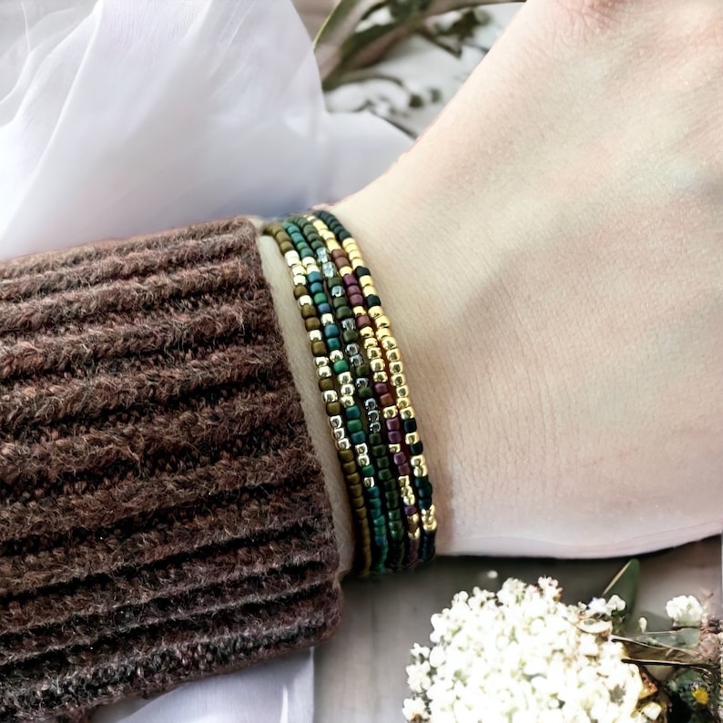 May include: A stack of colorful beaded bracelets in shades of green, gold, and brown. The bracelets are layered on a person's wrist.