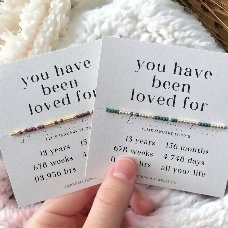 May include: Two cards with the text "you have been loved for" and a beaded bracelet on each card. The cards list the number of years, weeks, months, days, and hours a person has been loved. The text on the cards is "Ellie January 18, 20" and "Ellie January 18, 2019".