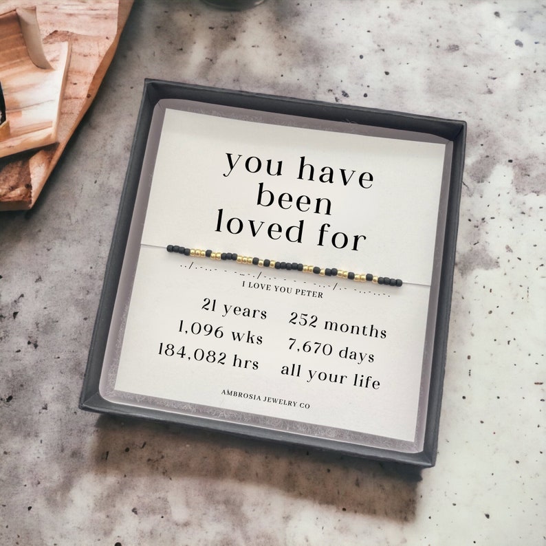 以下が含まれることがあります： 白いカードに「you have been loved for」と「I love you Peter」の文字が書かれた、黒と金のビーズのブレスレット。カードには、「21 years, 252 months, 1,096 wks, 7,670 days, 184,082 hrs, all your life」というテキストも記載されています。カードは黒い箱に入っています。