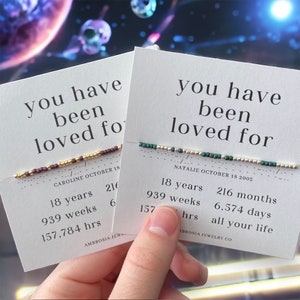 May include: Two cards with the text "you have been loved for" and the names Caroline and Natalie, along with their birthdates. The cards also list the number of years, weeks, months, days, and hours they have been alive. Each card has a beaded bracelet with different colored beads.