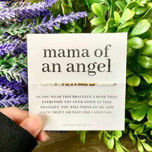 May include: A white card with the text "mama of an angel" printed in black. A white beaded bracelet with gold beads is on the card. The card also has the text "As you wear this bracelet, I hope that everytime you look down at this bracelet, you will think of me and know that I am safe and I love you." and "AMBROSIA JEWELRY CO" printed in black.