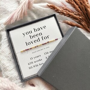 May include: A white card with the text "you have been loved for" and a beaded bracelet with gold and burgundy beads. The card also includes the date "ELLIE JANUARY 18, 2019" and a list of time measurements: "13 years", "156 mo", "678 weeks", "4,7", "113,956 hrs".