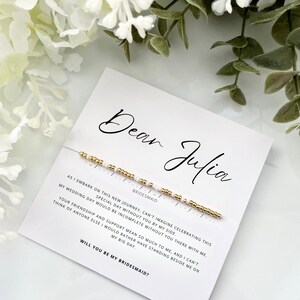 May include: A delicate bracelet featuring gold and white beads arranged in a Morse code pattern, attached to a white card with the text Dear Julia, the word BRIDESMAID, and a printed message asking the recipient to be a bridesmaid.