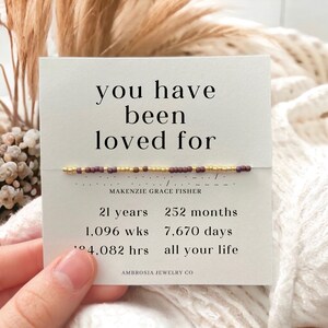 K&ouml;nnte beinhalten: Eine wei&szlig;e Karte mit dem Text "you have been loved for" und einem Perlenarmband mit goldenen und kastanienbraunen Perlen. Die Karte enth&auml;lt auch den Namen "MAKENZIE GRACE FISHER" und den Text "21 Jahre, 252 Monate, 1.096 Wochen, 7.670 Tage, 24.082 Stunden, dein ganzes Leben" und "AMBROSIA JEWELRY CO."