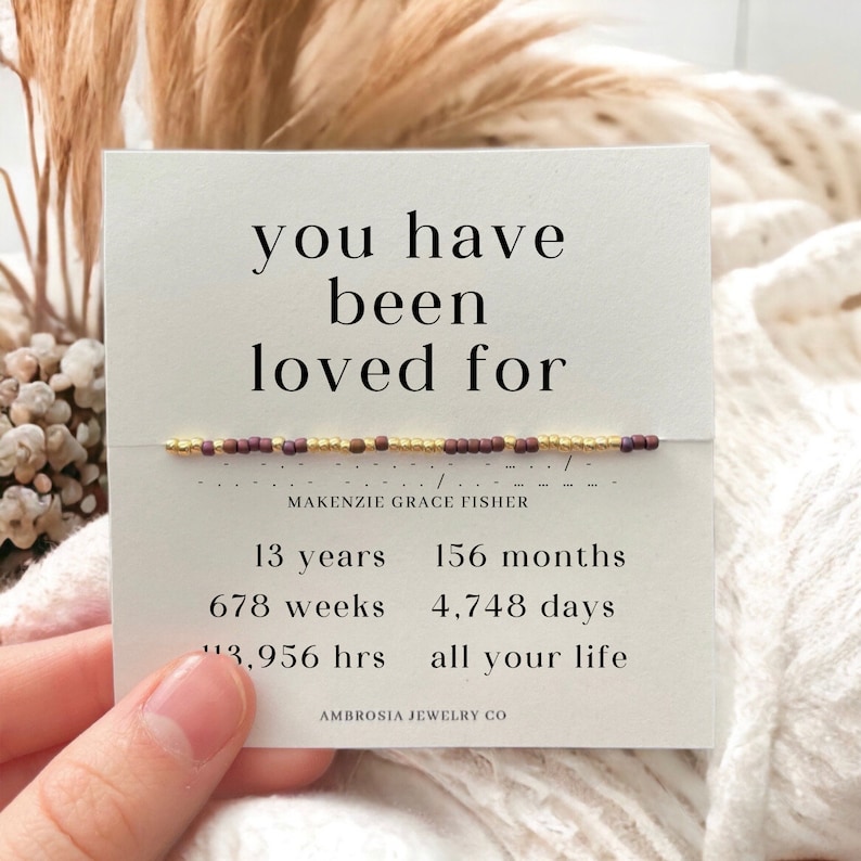 May include: A white card with the text "you have been loved for" and a beaded bracelet with gold and brown beads. The card also includes the name "MAKENZIE GRACE FISHER" and the text "13 years, 156 months, 678 weeks, 4,748 days, 13,956 hrs, all your life" and "AMBROSIA JEWELRY CO."