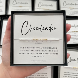 May include: White and gold beaded bracelet with the word "Cheerleader" and the name "Katelyn" on a white card with the text "The greatness of a cheerleader isn't determined by how high she jumps, but by the boundless spirit she brings." and the text "Ambrosia Jewelry Co."