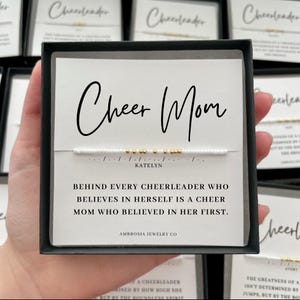 May include: A black box with a white card that reads "Cheer Mom" in script. A white beaded bracelet with gold beads is visible. The card also says "Behind every cheerleader who believes in herself is a cheer mom who believed in her first."