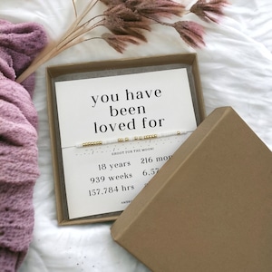 May include: A white and gold beaded bracelet with the text "you have been loved for" and "SHOOT FOR THE MOON!" on a white card in a brown box. The card also lists the time in years, weeks, and hours.