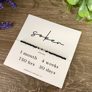 May include: Black beaded bracelet with silver beads and the word "sober" written on a white card. The card also includes the text "1 month, 730 hrs, 4 weeks, 30 days" and the company name "AMBROISA JEWELRY CO."