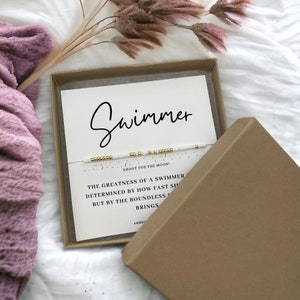 May include: A white box with a white and gold beaded bracelet inside. The box has a white card with the text "Swimmer" and a quote about the greatness of a swimmer. "Shoot for the moon!"