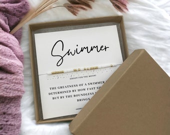 May include: A white box with a white and gold beaded bracelet inside. The box has a white card with the text "Swimmer" and a quote about the greatness of a swimmer. "Shoot for the moon!"