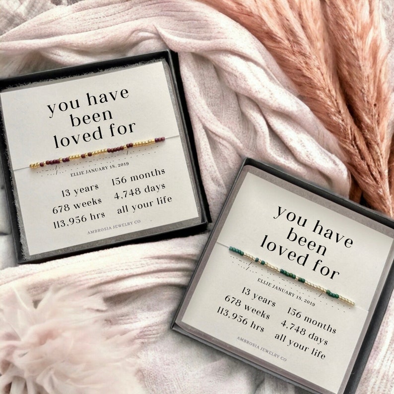 May include: Two small black boxes with white text that reads "you have been loved for" and a beaded bracelet inside. The text also includes the date "Ellie January 15, 2019" and a list of time units: 13 years, 156 months, 678 weeks, 4,748 days, 113,956 hours, and "all your life". The boxes are on a pink and white fuzzy surface.