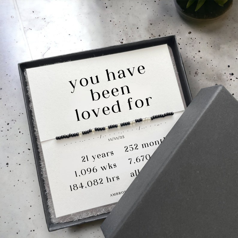 以下が含まれることがあります： 黒い文字で「you have been loved for」と書かれた白いカードと、黒と白のビーズでできたブレスレット。カードには「21 years, 1,096 wks, 184,082 hrs, 11/11/23, 252 mont, 7,670, all」というテキストも含まれています。