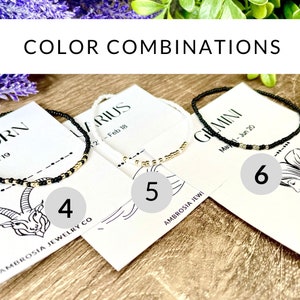 May include: Four different zodiac sign bracelets with black, white, and gold beads. The bracelets are displayed on cards with the zodiac sign name and dates. The cards are on a wooden surface.
