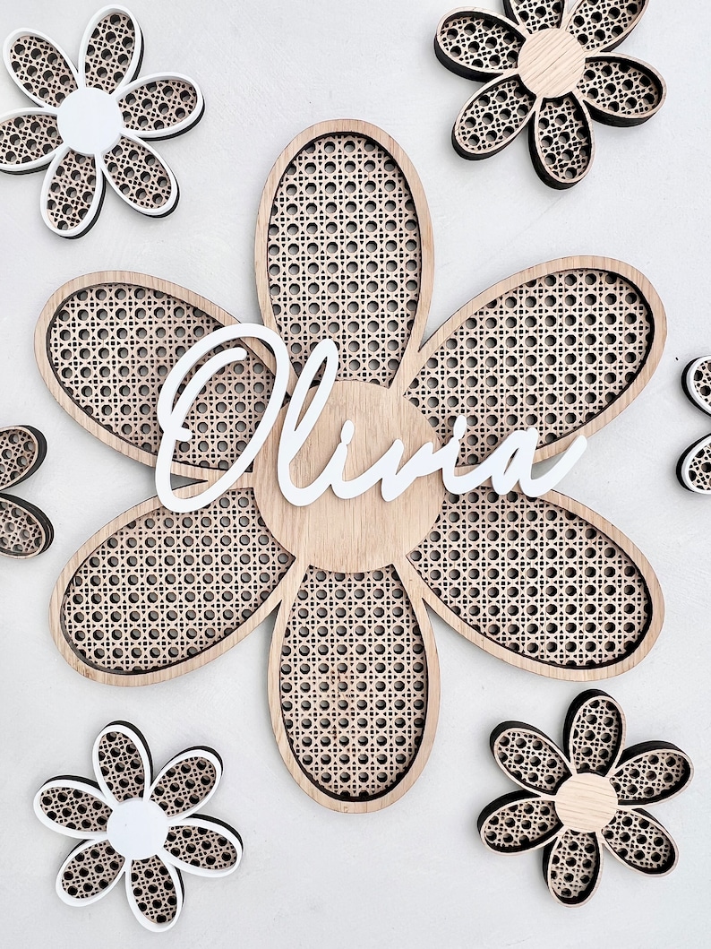 K&ouml;nnte beinhalten: Eine Holz-Wanddekoration mit einer gro&szlig;en, sechszackigen Blume mit einem gewebten Muster. Die Blume tr&auml;gt den Namen "Olivia" in wei&szlig;er Schrift. F&uuml;nf kleinere, sechszackige Blumen umgeben die gro&szlig;e Blume, von denen zwei wei&szlig; und drei braun sind.