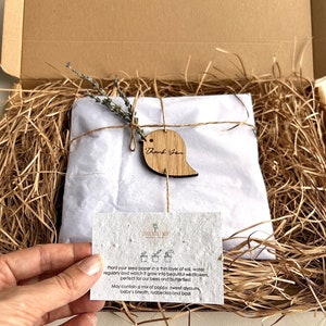 K&ouml;nnte beinhalten: Ein brauner Karton, gef&uuml;llt mit braunem, zerkleinertem Papier. Ein wei&szlig;er Umschlag mit einem kleinen Lavendelzweig ist mit Schnur und einem Holzschild mit der Aufschrift "Danke" gebunden. Eine kleine Karte mit Anweisungen zum Pflanzen von Saatpapier ist sichtbar. Die Karte sagt: "Pflanzen Sie Ihr Saatpapier in eine d&uuml;nne Schicht Erde, gie&szlig;en Sie es regelm&auml;&szlig;ig und beobachten Sie, wie es zu wundersch&ouml;nen Wildblumen heranw&auml;chst, perfekt f&uuml;r unsere Bienen und Schmetterlinge! Kann eine Mischung aus Mohn, s&uuml;&szlig;em Alyssum, Babyschnabel, Rudbeckie und Basilikum enthalten."
