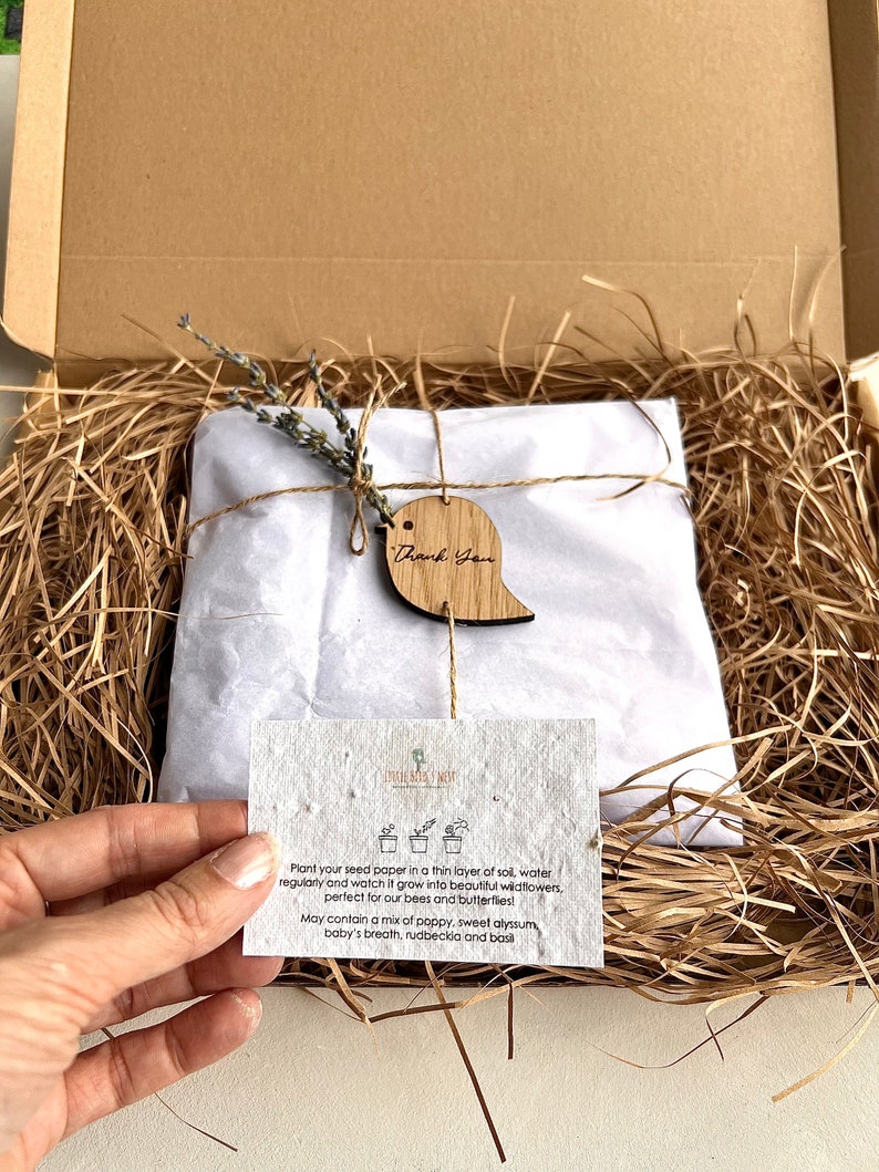 K&ouml;nnte beinhalten: Ein brauner Karton, gef&uuml;llt mit braunem, zerkleinertem Papier. Ein wei&szlig;er Umschlag mit einem kleinen Lavendelzweig ist mit Schnur und einem Holzschild mit der Aufschrift "Danke" gebunden. Eine kleine Karte mit Anweisungen zum Pflanzen von Saatpapier ist sichtbar. Die Karte sagt: "Pflanzen Sie Ihr Saatpapier in eine d&uuml;nne Schicht Erde, gie&szlig;en Sie es regelm&auml;&szlig;ig und beobachten Sie, wie es zu wundersch&ouml;nen Wildblumen heranw&auml;chst, perfekt f&uuml;r unsere Bienen und Schmetterlinge! Kann eine Mischung aus Mohn, s&uuml;&szlig;em Alyssum, Babyschnabel, Rudbeckie und Basilikum enthalten."