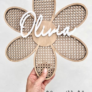 K&ouml;nnte beinhalten: Eine holzerne, blumenf&ouml;rmige Wanddekoration mit einem geflochtenen Rohrgeflecht. Der Name "Olivia" ist in Wei&szlig; in der Mitte der Blume geschrieben. Die Blume hat einen Durchmesser von 30 cm.