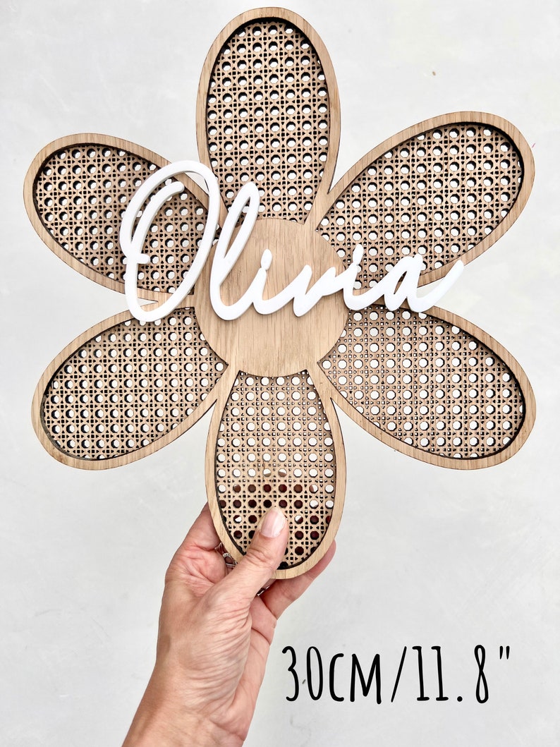 K&ouml;nnte beinhalten: Eine holzerne, blumenf&ouml;rmige Wanddekoration mit einem geflochtenen Rohrgeflecht. Der Name "Olivia" ist in Wei&szlig; in der Mitte der Blume geschrieben. Die Blume hat einen Durchmesser von 30 cm.