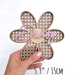 K&ouml;nnte beinhalten: Ein h&ouml;lzernes Schild in Form einer Blume mit einem gewebten Muster und dem Namen "Daisy" in Pink darauf. Das Schild hat einen Durchmesser von 15 cm und ist in einem matten Rosaton lackiert.